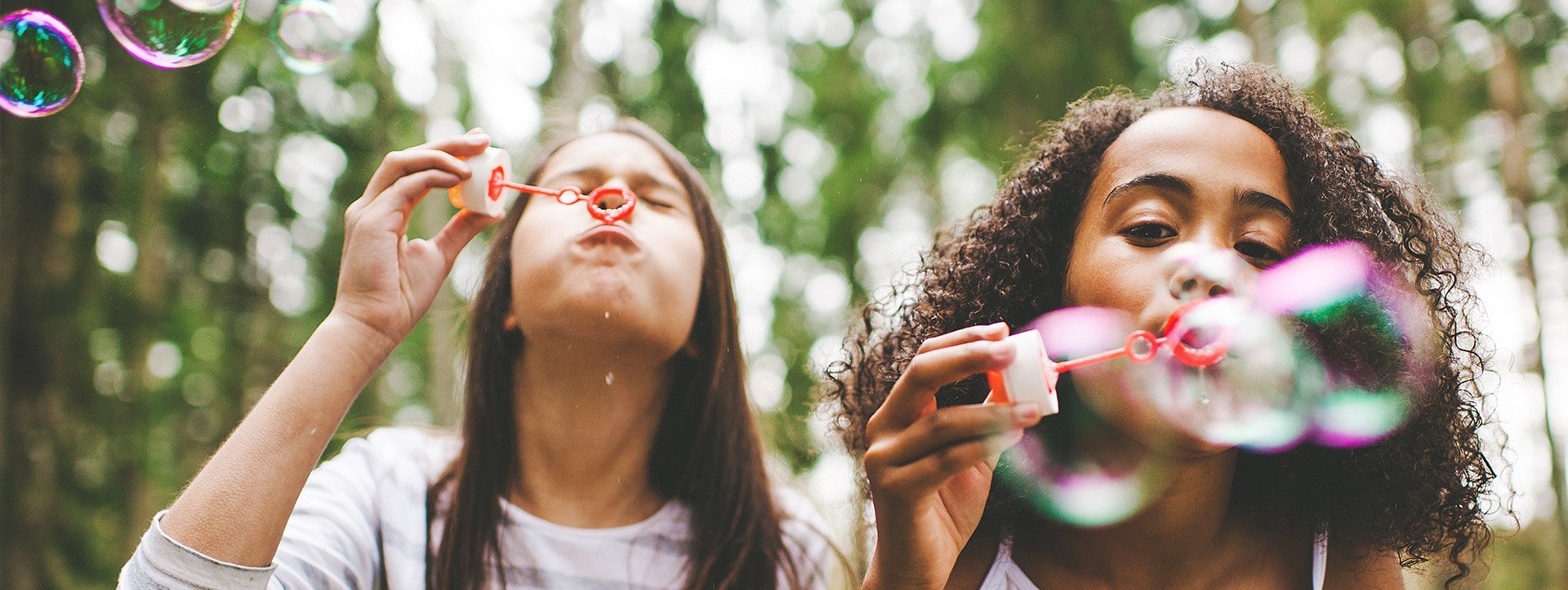 two children blow bubbles through bubble wands toward the camera, trees in background